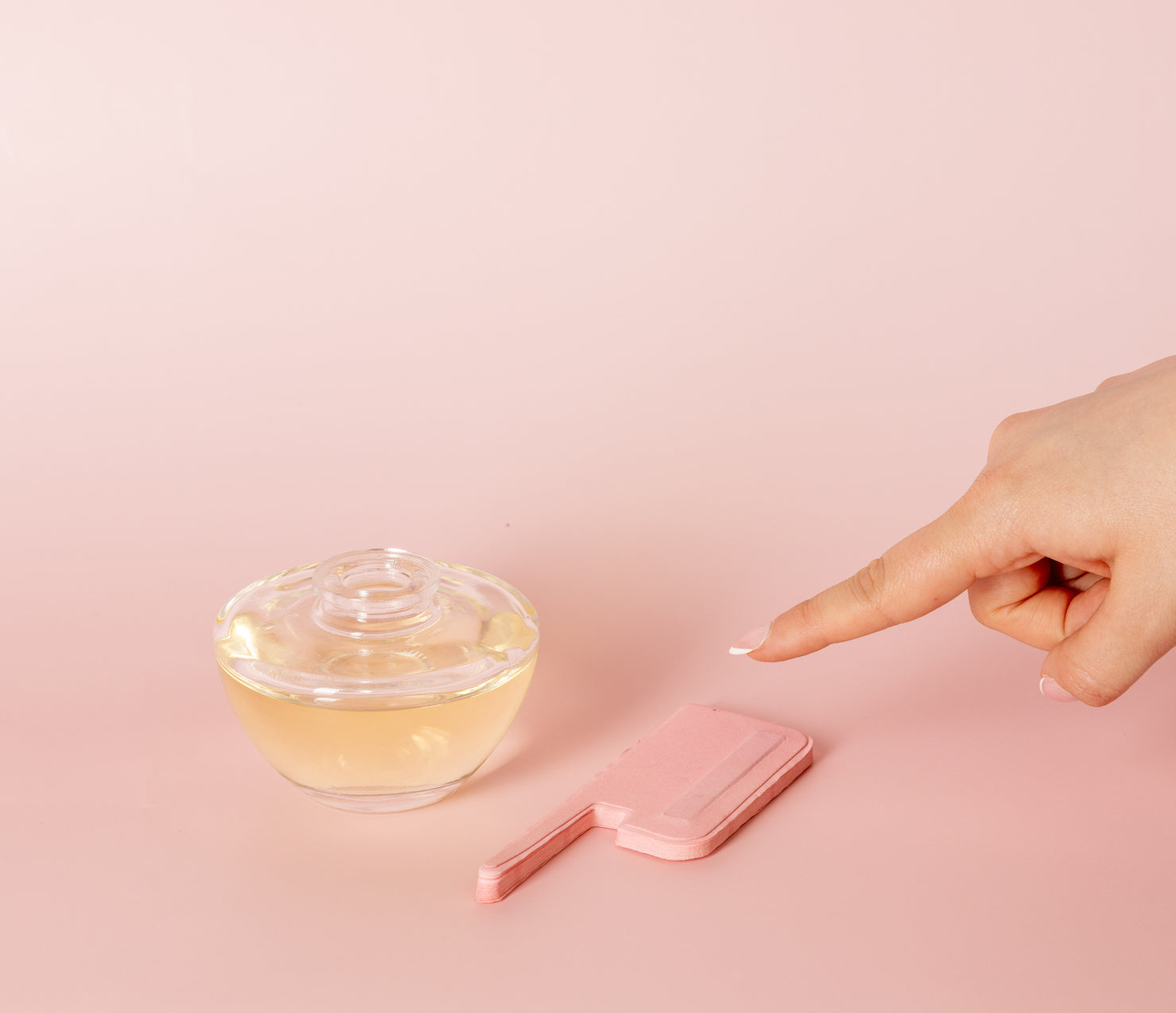 A hand points toward a clear glass container filled with liquid and a pink wax spatula on a light pink background.