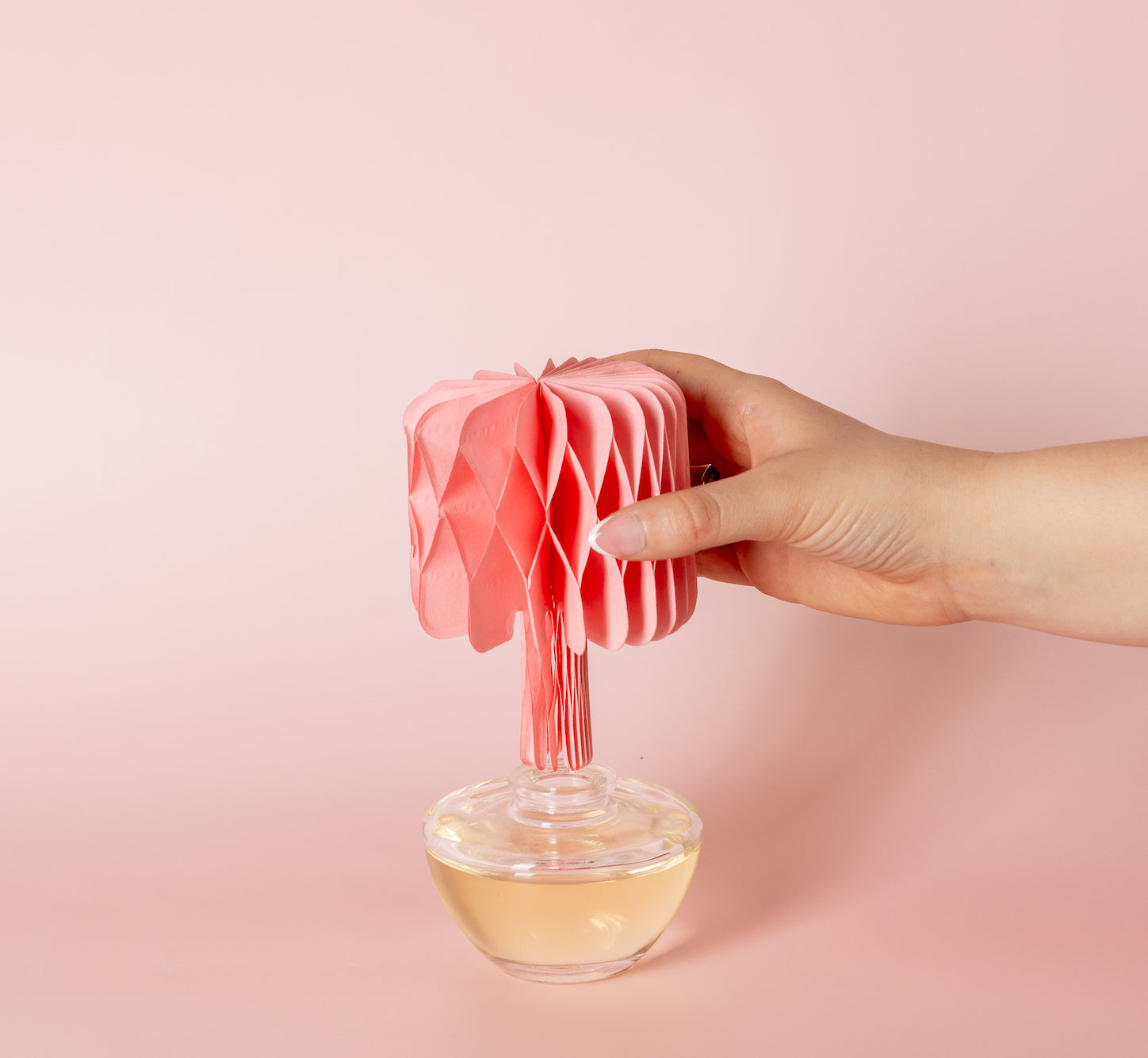 A hand holds a pink paper decoration resembling a tree, which is attached to a round glass container filled with yellow liquid, against a light pink background.