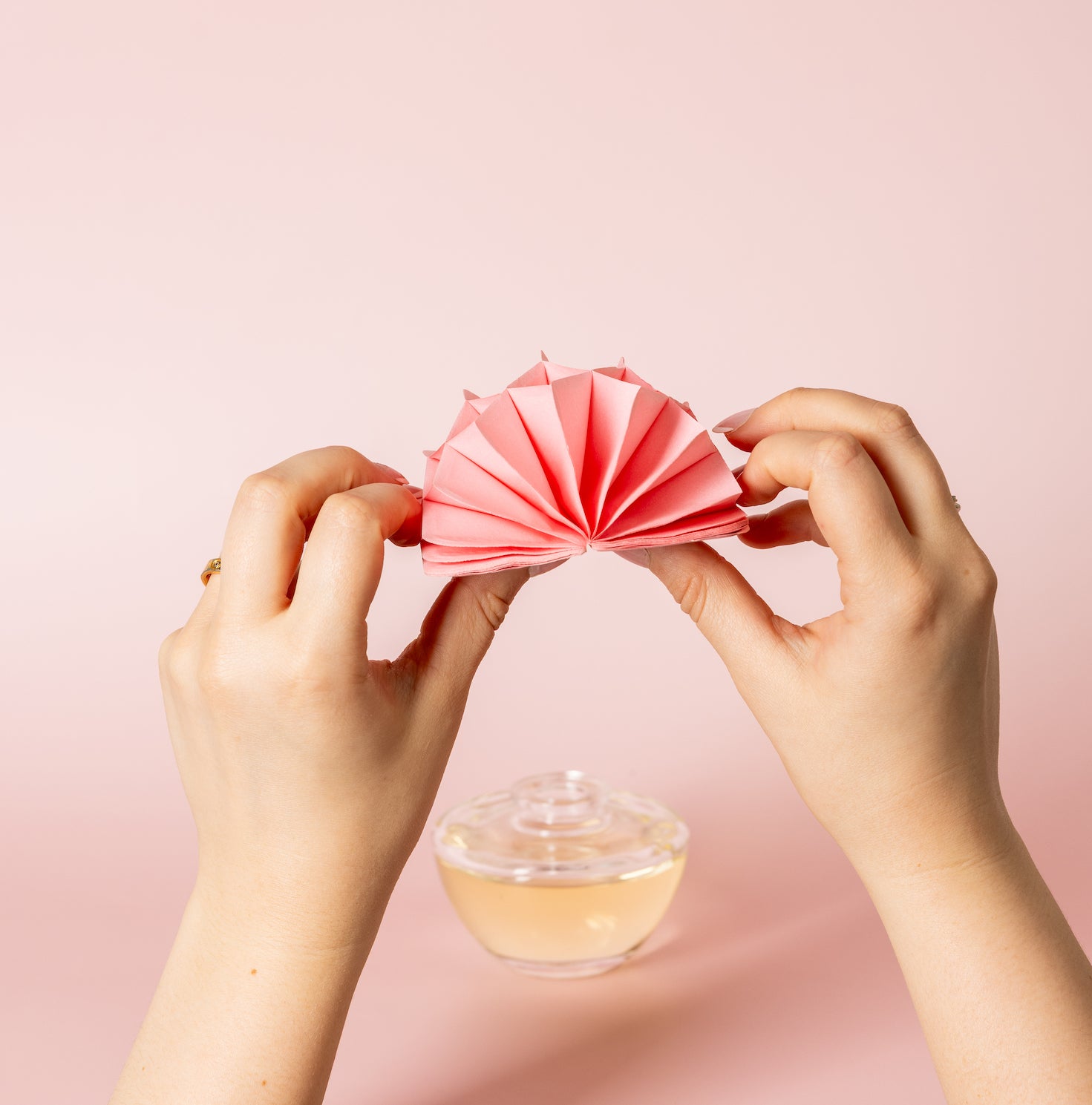 Two hands folding a pink paper fan on a light pink surface, with a round glass bottle containing yellow liquid in the background.