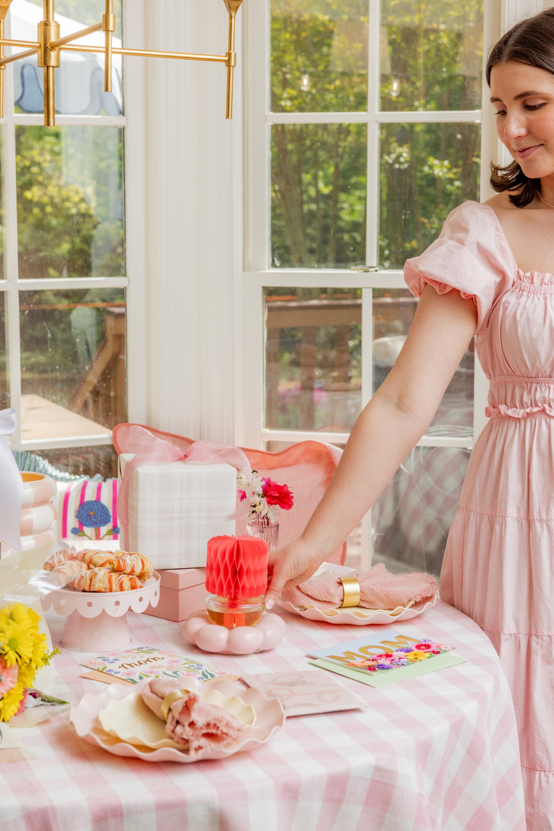 A woman in a pink dress arranges napkins on a pastel-themed party table with cookies, flowers, and presents, in a sunny room with large windows.