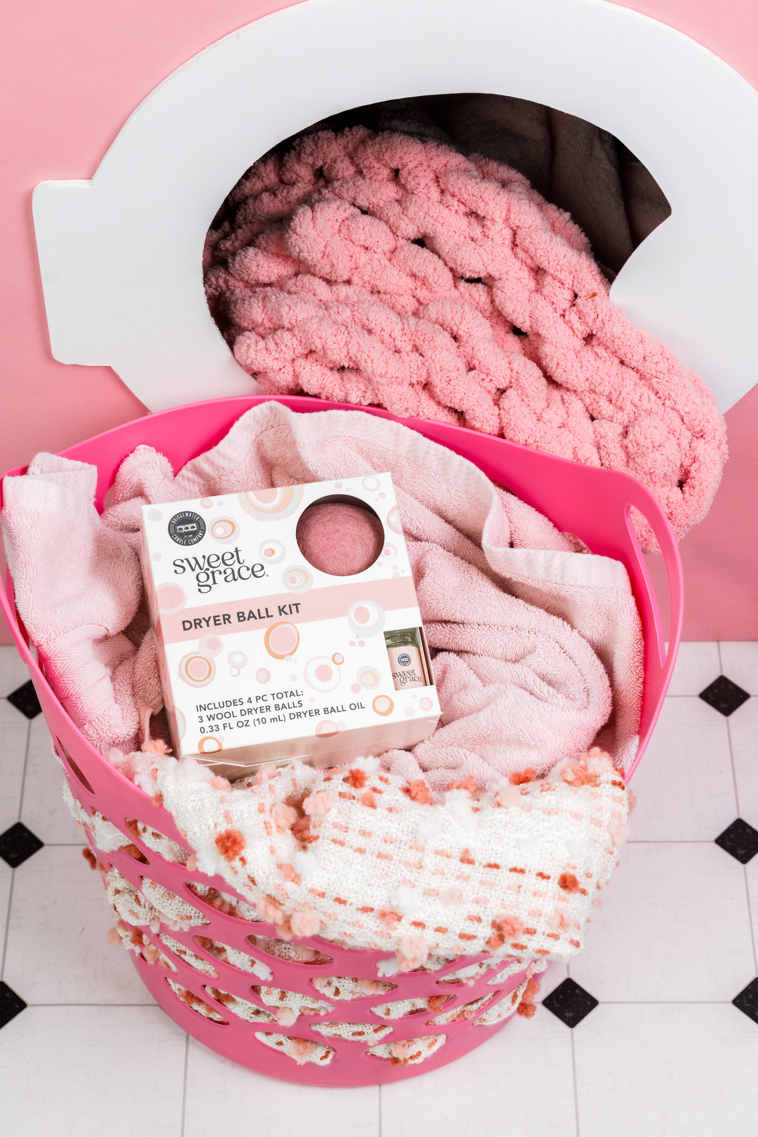 A pink laundry basket filled with pink towels and a Sweet Grace dryer ball kit sits in front of a dryer with a pink blanket inside.