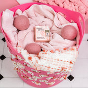 A pink laundry basket holds light pink towels, wool dryer balls, a pink and white blanket, and a Sweet Grace Dryer Ball Refill by Bridgewater Candle for added fragrance on a white tiled floor.