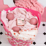 The Bridgewater Candle Sweet Grace Dryer Ball Kit, including pink towels, wool dryer balls, and Sweet Grace Dryer Ball Oil, sits in a pink laundry basket beside a chunky knit blanket—perfect for fragranced laundry.