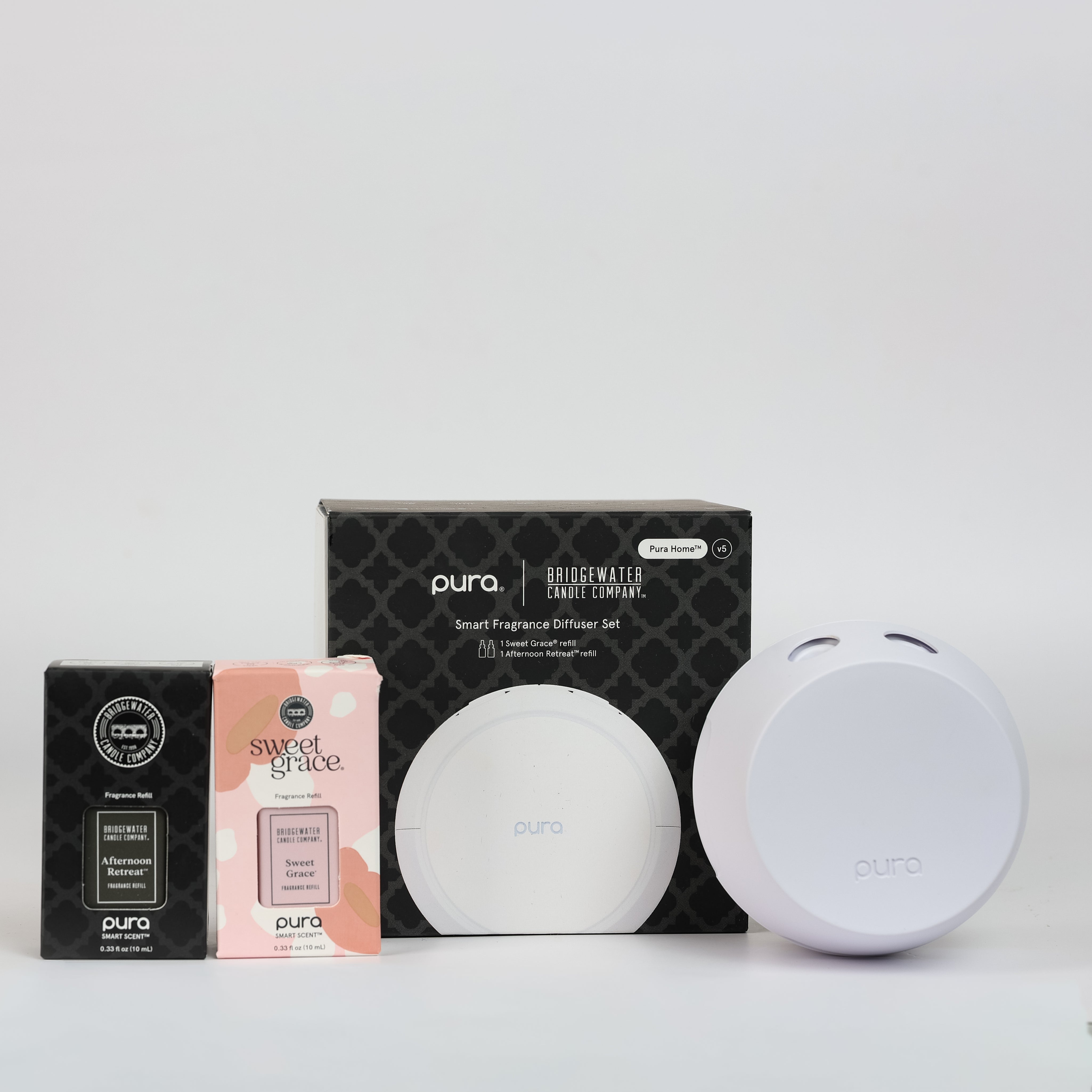 The PURA + Bridgewater Smart Home Diffuser Set with Sweet Grace and Afternoon Retreat V5 by Bridgewater Candle features a round white PURA diffuser, sleek black packaging, and fragrance boxes for Sweet Grace and Afternoon Retreat, all displayed on a white background.