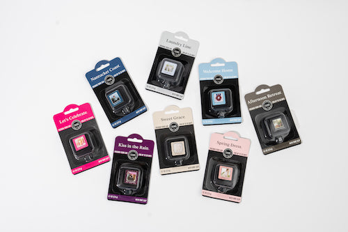 Nine air fresheners in individual plastic packaging are arranged on a white surface. Each package is labeled with a different scent and color, including pink, blue, purple, gray, and black themes.