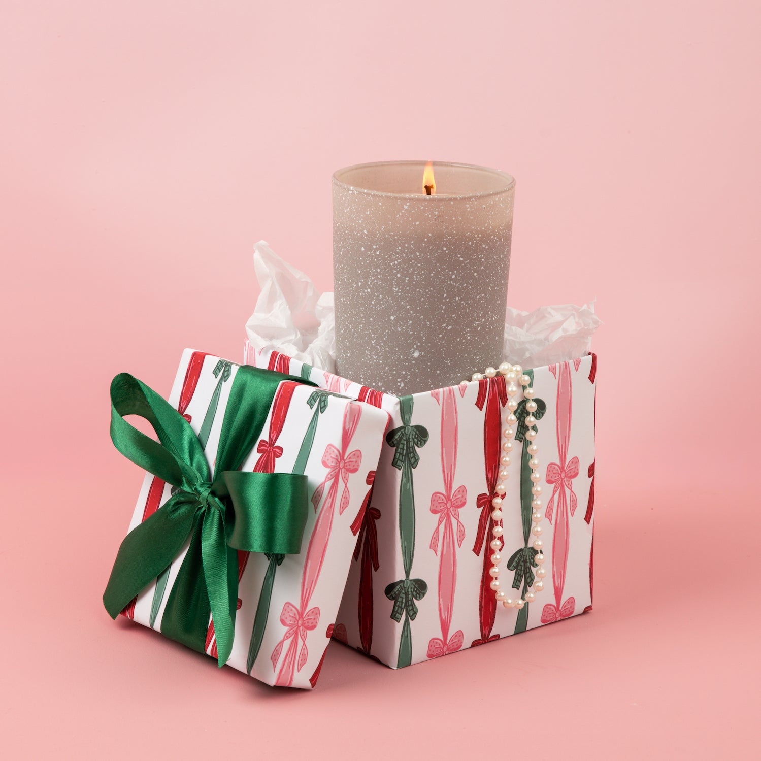 A Sweet Grace Frosted Gray Candle by Bridgewater Candle burns in an open gift box with a green ribbon, pearl bracelet, and the Light A Candle Feed A Child message, resting on a pink background.