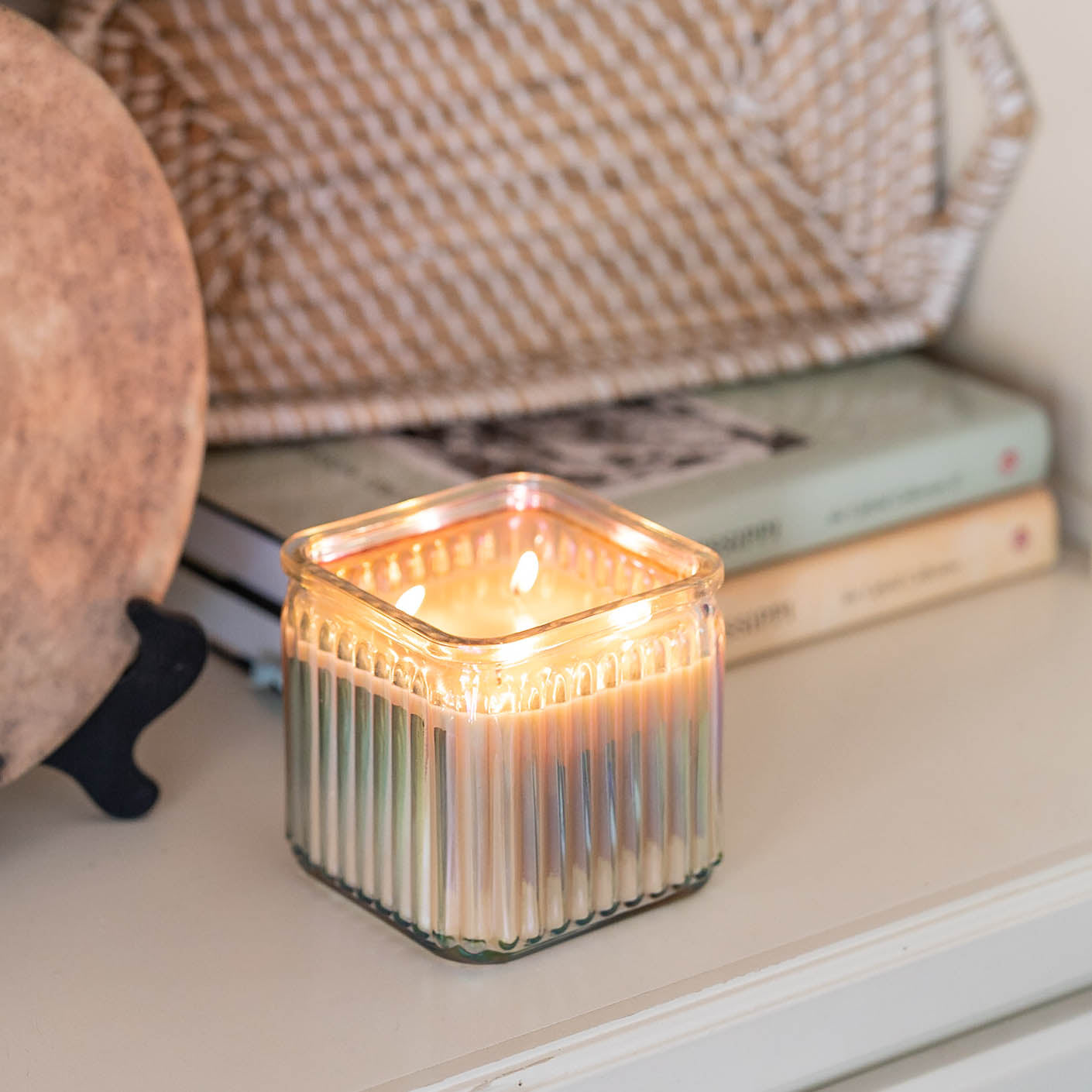 A lit candle in a square, ribbed glass container sits on a shelf near stacked books and a decorative woven tray. The warm flame creates a cozy ambiance.