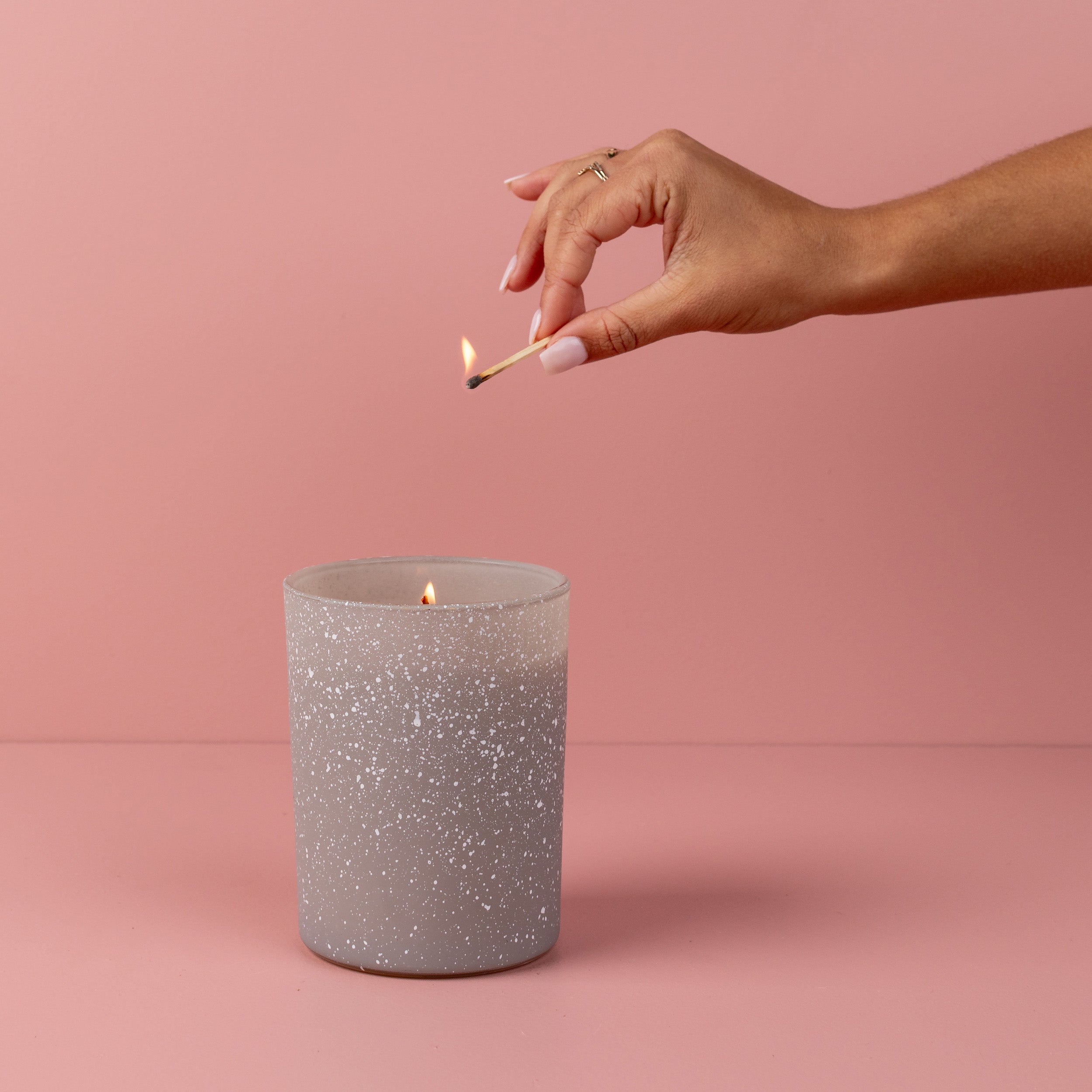 A hand with manicured nails holds a lit match above a Bridgewater Candle Sweet Grace Frosted Gray Candle in a glass jar, set on a pink surface. Ideal as a holiday gift to Light A Candle Feed A Child.