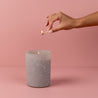 A hand with manicured nails holds a lit match above a Bridgewater Candle Sweet Grace Frosted Gray Candle in a glass jar, set on a pink surface. Ideal as a holiday gift to Light A Candle Feed A Child.