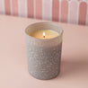 A lit Bridgewater Candle Sweet Grace Frosted Gray Candle sits on a pale pink surface with a pink and white patterned wall—an ideal holiday gift that supports Light A Candle Feed A Child.