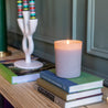 A lit Bridgewater Candle Sweet Grace Frosted Gray Candle rests on books atop a wooden surface, with a white holder and striped candles in the background—a cozy, inviting scene that makes an ideal holiday gift.