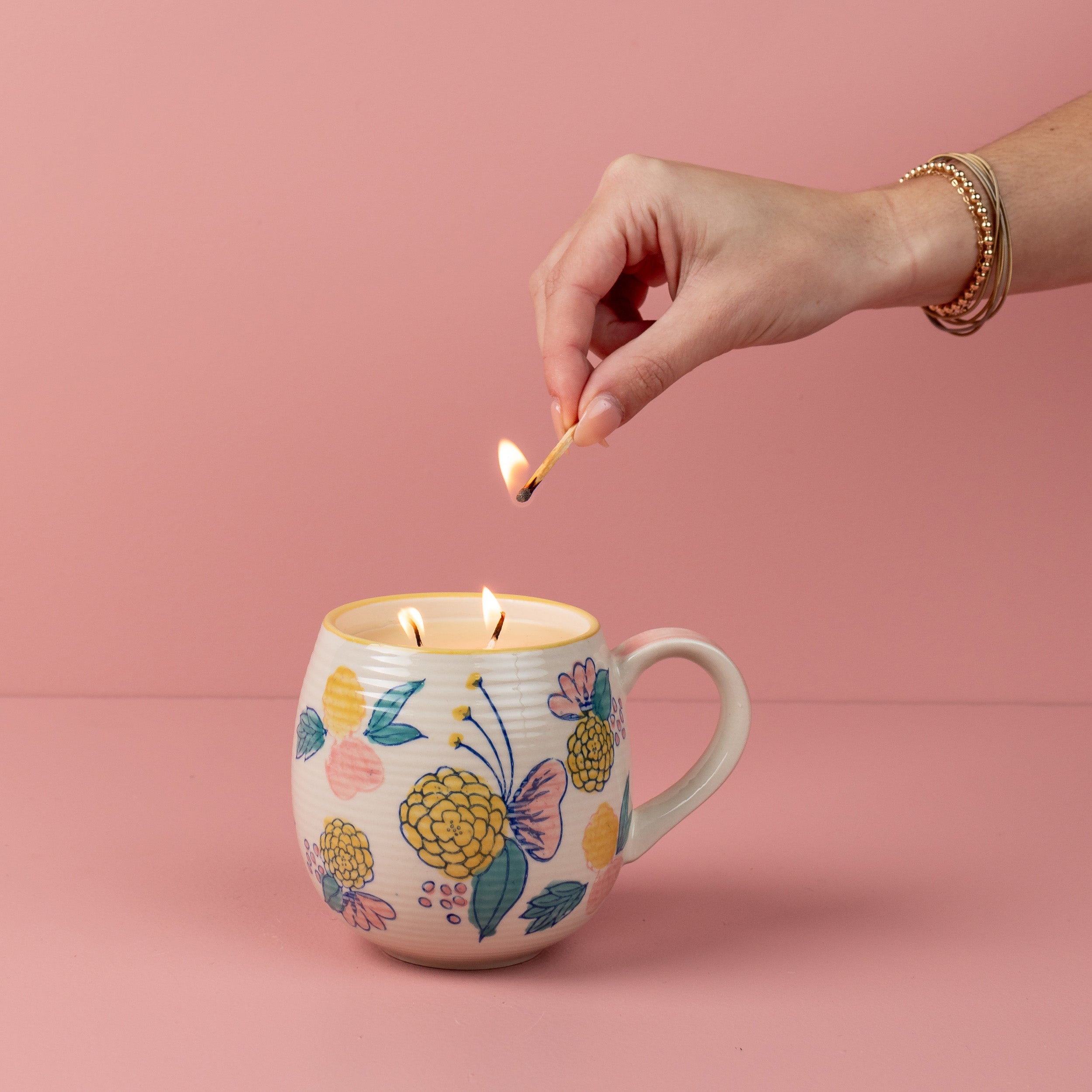 A hand lights the Bridgewater Candle Sweet Grace Mug Candle, placed inside a blush floral mug against a pink background.