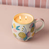A lit Bridgewater Candle Sweet Grace Mug Candle with two flames sits in a blush floral mug adorned with vibrant flowers on a light pink surface, set against a blurred pink and white striped background.