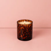 A Sweet Grace Collection Candle #064 by Bridgewater Candle Company, featuring soy-blend wax in a brown tortoise glass holder, is shown on a pink surface with a matching pink background.
