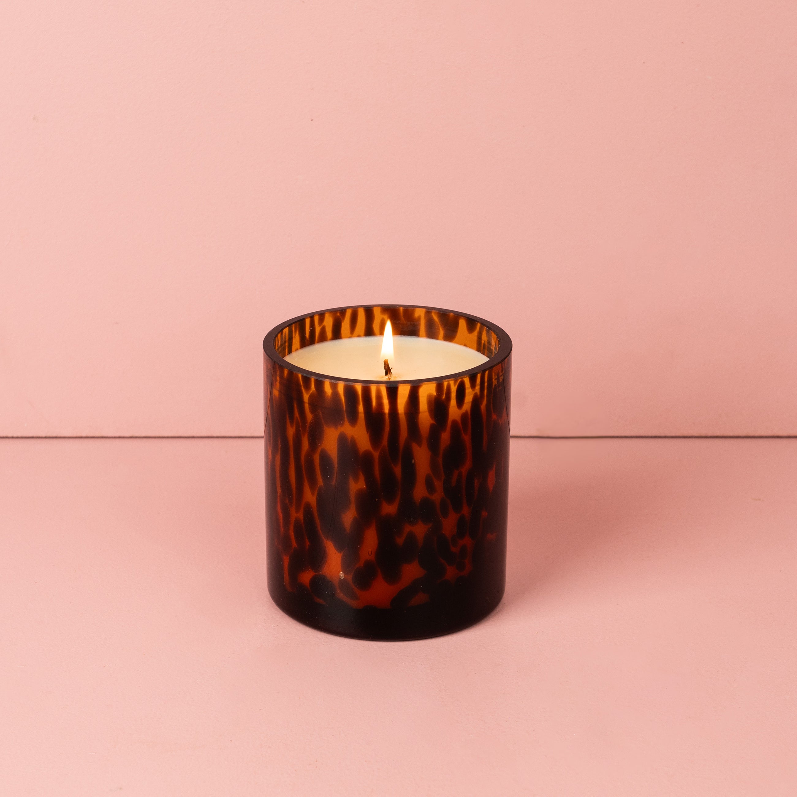 A Sweet Grace Collection Candle #064 by Bridgewater Candle Company, featuring soy-blend wax in a brown tortoise glass holder, is shown on a pink surface with a matching pink background.