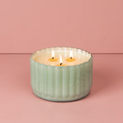 The Sweet Grace Collection Candle #065 by Bridgewater Candle Company features a mint green ribbed ceramic holder filled with soy-blend wax and three lit wicks, displayed against a pink background.