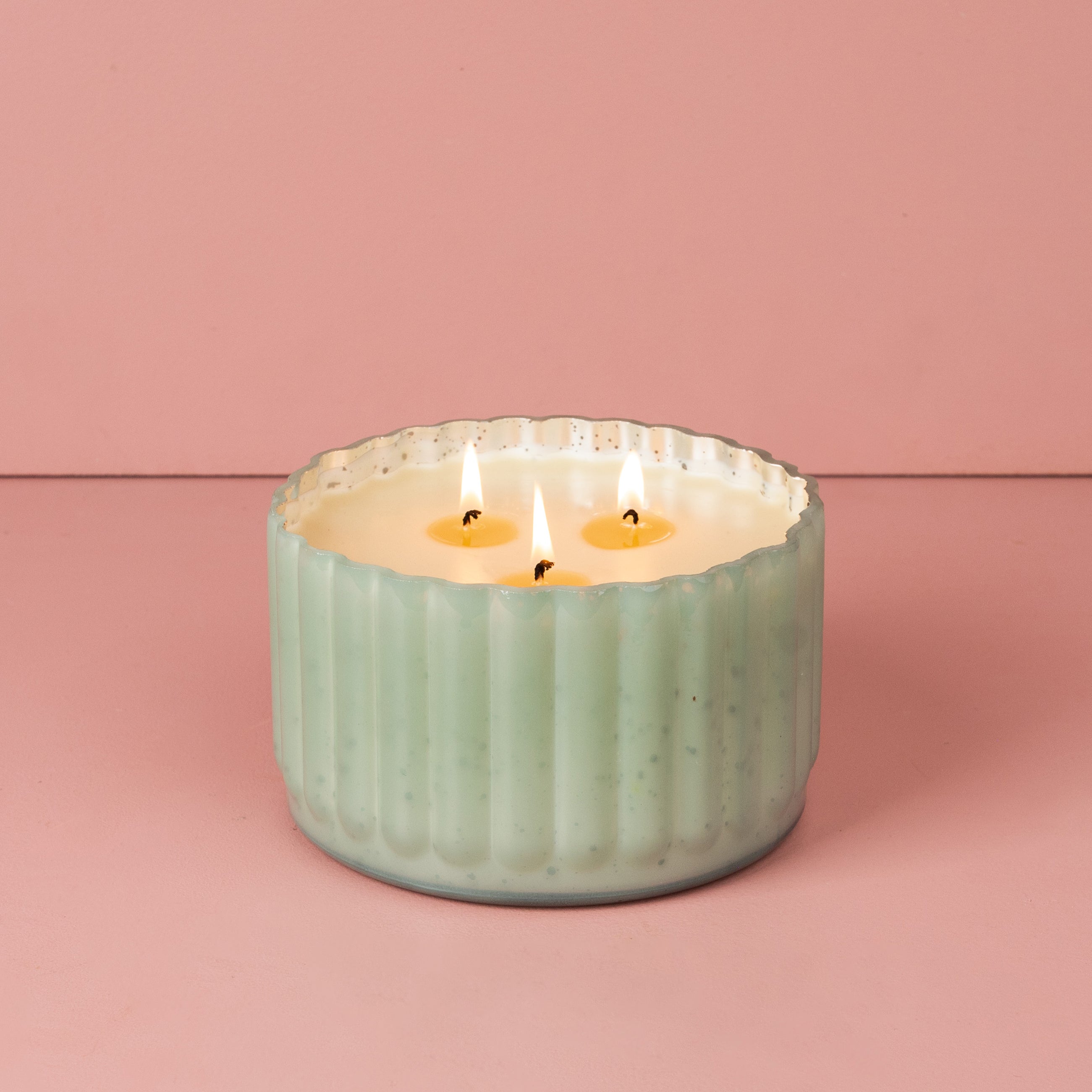 The Sweet Grace Collection Candle #065 by Bridgewater Candle Company features a mint green ribbed ceramic holder filled with soy-blend wax and three lit wicks, displayed against a pink background.