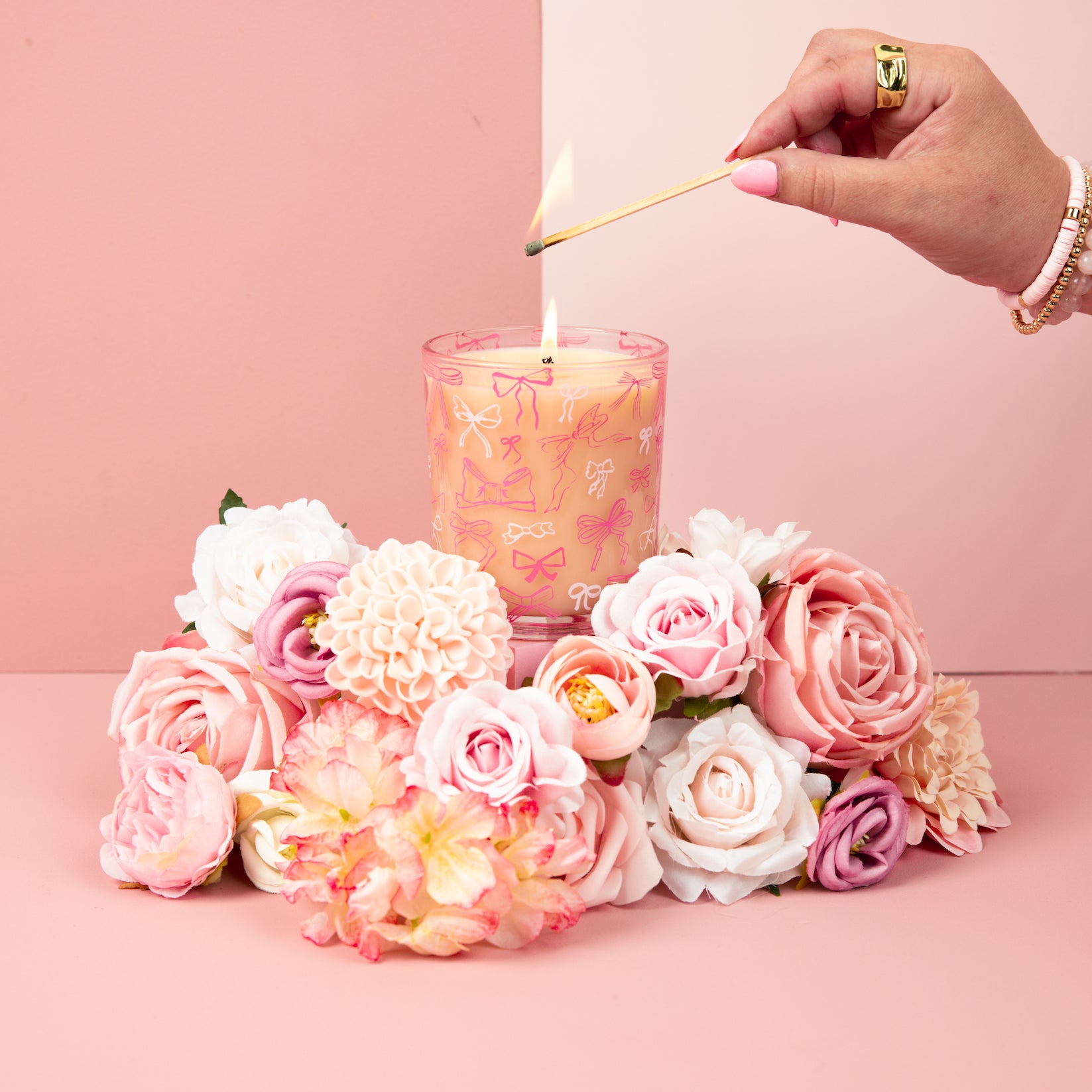 A hand lights the Sweet Grace Pink Bow Candle by Bridgewater Candle, crafted from soy-blend wax and adorned with pink bows. The candle sits among pink and white flowers on a soft pink background.