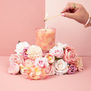 A hand lights the Sweet Grace Pink Bow Candle by Bridgewater Candle, crafted from soy-blend wax and adorned with pink bows. The candle sits among pink and white flowers on a soft pink background.