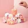 A hand lights the Sweet Grace Pink Bow Candle by Bridgewater Candle, crafted from soy-blend wax and adorned with pink bows. The candle sits among pink and white flowers on a soft pink background.
