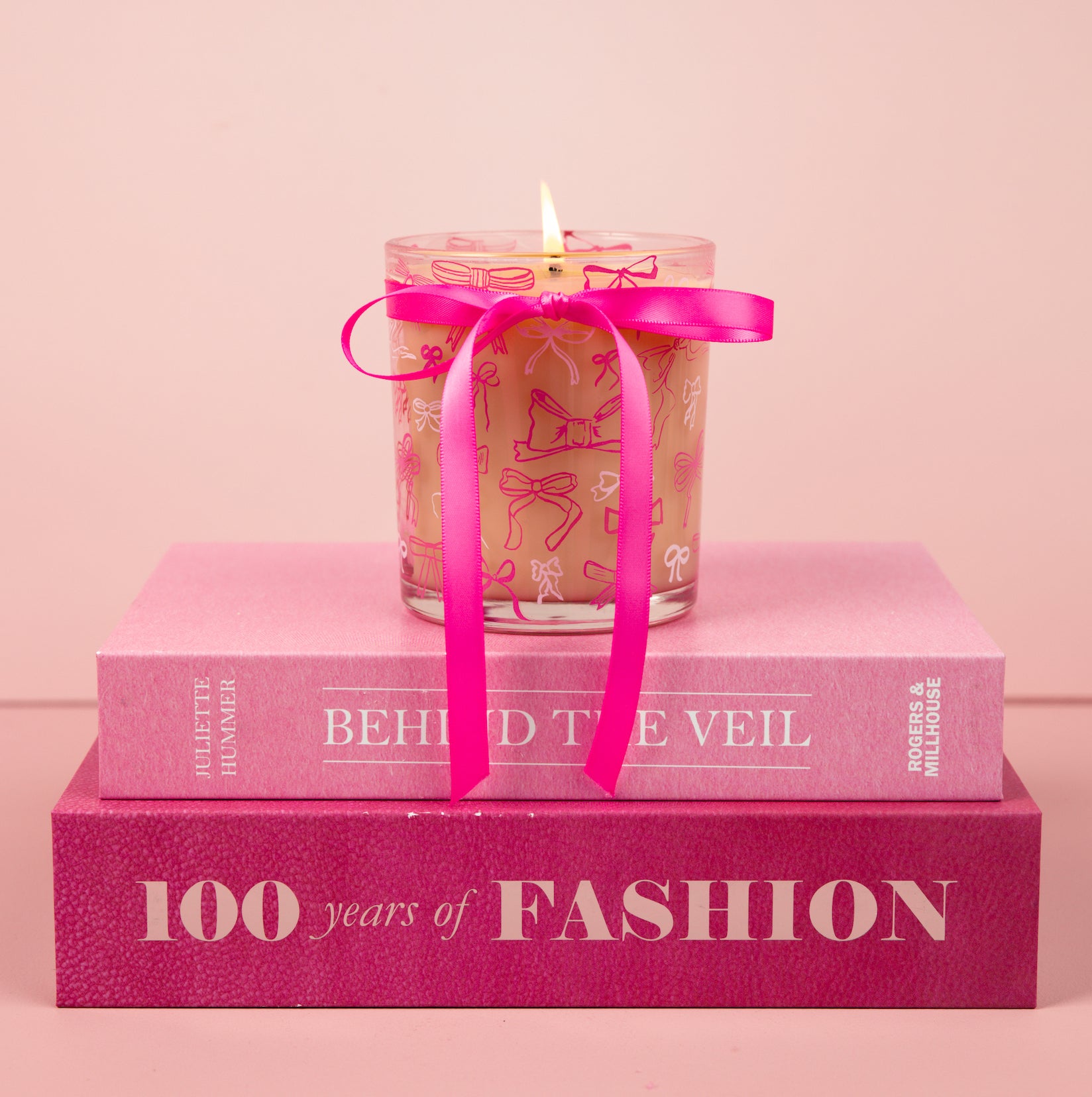 The Bridgewater Candle Sweet Grace Pink Bow Candle, made from soy-blend wax, sits atop two stacked pink books—BEHIND THE VEIL and 100 Years of FASHION—against a soft pink background.