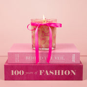 The Bridgewater Candle Sweet Grace Pink Bow Candle, made from soy-blend wax, sits atop two stacked pink books—BEHIND THE VEIL and 100 Years of FASHION—against a soft pink background.