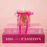 The Bridgewater Candle Sweet Grace Pink Bow Candle, made from soy-blend wax, sits atop two stacked pink books—BEHIND THE VEIL and 100 Years of FASHION—against a soft pink background.