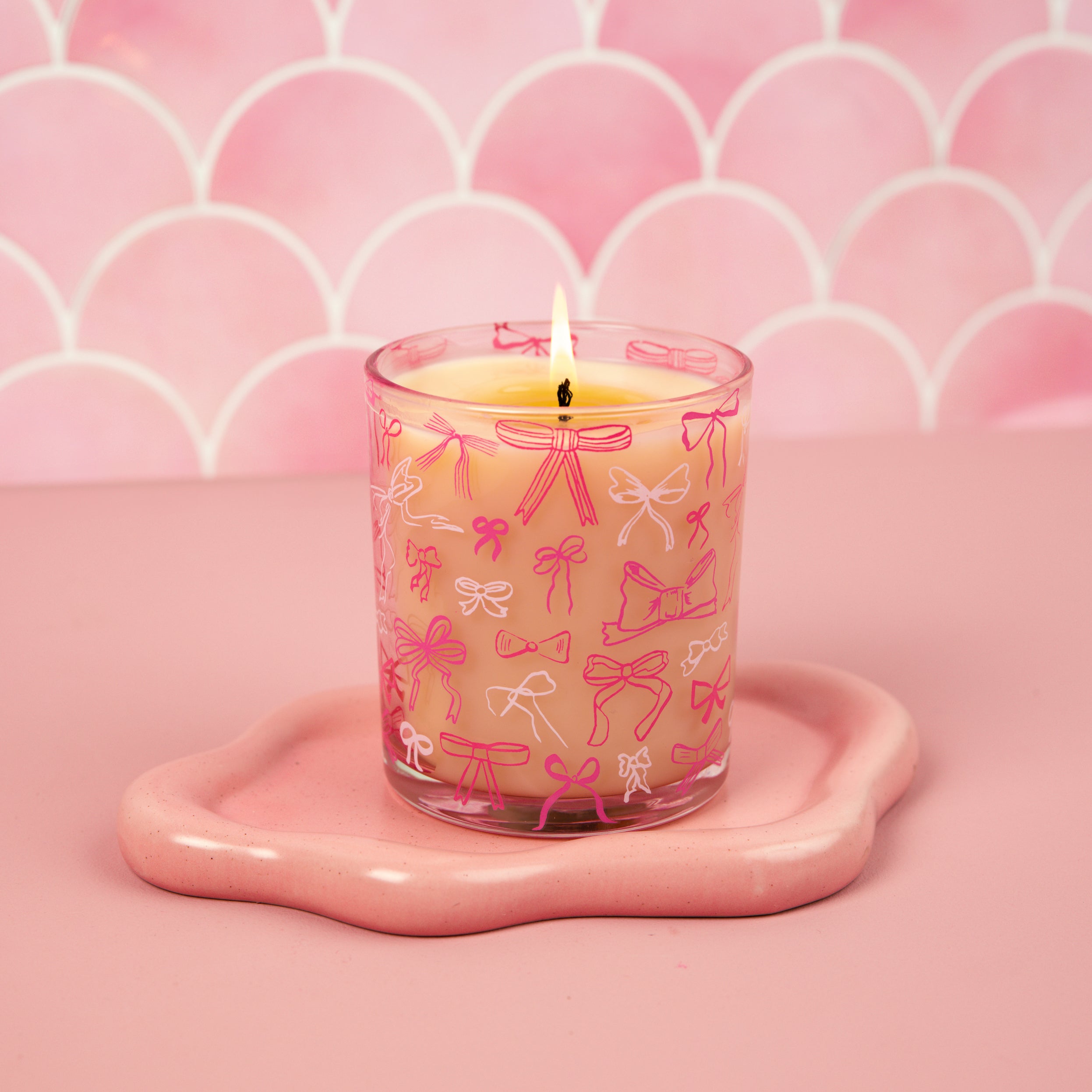 The Bridgewater Candle Sweet Grace Pink Bow Candle, crafted from soy-blend wax, glows in a glass holder with pink bows on a wavy-edged pink tray against a scalloped pink background.