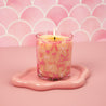 The Bridgewater Candle Sweet Grace Pink Bow Candle, crafted from soy-blend wax, glows in a glass holder with pink bows on a wavy-edged pink tray against a scalloped pink background.