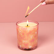 A hand with pink-painted nails lights the Bridgewater Candle Sweet Grace Pink Bow Candle, crafted from soy-blend wax in a glass decorated with bows, set against a pink background.