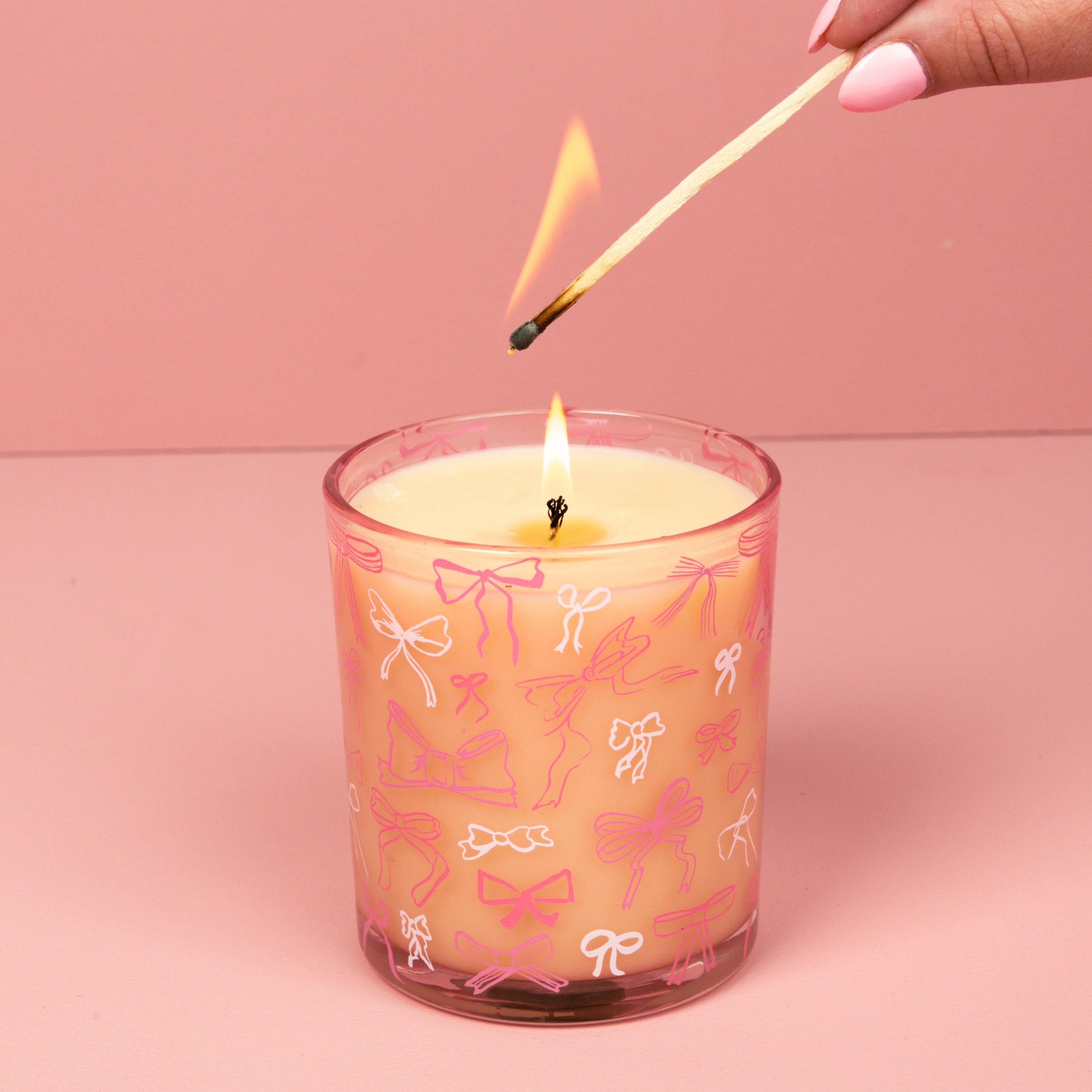 A hand with pink-painted nails lights the Bridgewater Candle Sweet Grace Pink Bow Candle, crafted from soy-blend wax in a glass decorated with bows, set against a pink background.
