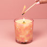 A hand with pink-painted nails lights the Bridgewater Candle Sweet Grace Pink Bow Candle, crafted from soy-blend wax in a glass decorated with bows, set against a pink background.