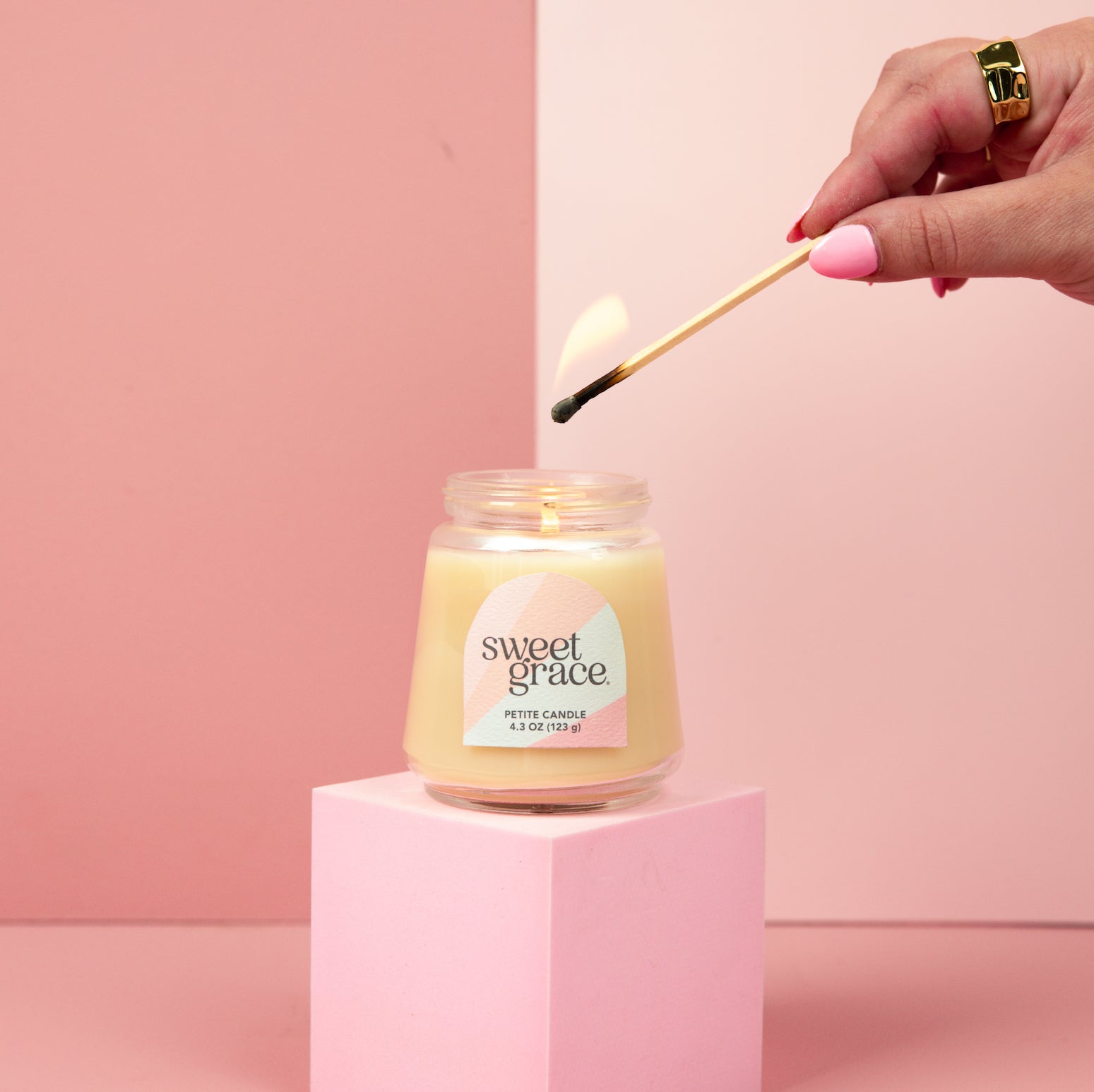 A hand with pink nail polish lights the Bridgewater Candle Sweet Grace Collection Candle #078—crafted from soy-blend wax—in a petite glass jar. The candle sits on a pink pedestal against a pink and cream backdrop.
