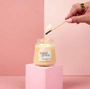 A hand with pink nail polish lights the Bridgewater Candle Sweet Grace Collection Candle #078—crafted from soy-blend wax—in a petite glass jar. The candle sits on a pink pedestal against a pink and cream backdrop.