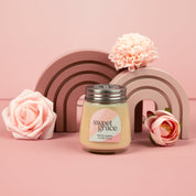 The Bridgewater Candle Sweet Grace Collection Candle #078, a soy-blend wax candle in a petite glass jar, rests on a pink surface with pink artificial flowers and decorative rainbow arches, creating a charming scene against a pink backdrop.