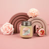 The Bridgewater Candle Sweet Grace Collection Candle #078, a soy-blend wax candle in a petite glass jar, rests on a pink surface with pink artificial flowers and decorative rainbow arches, creating a charming scene against a pink backdrop.