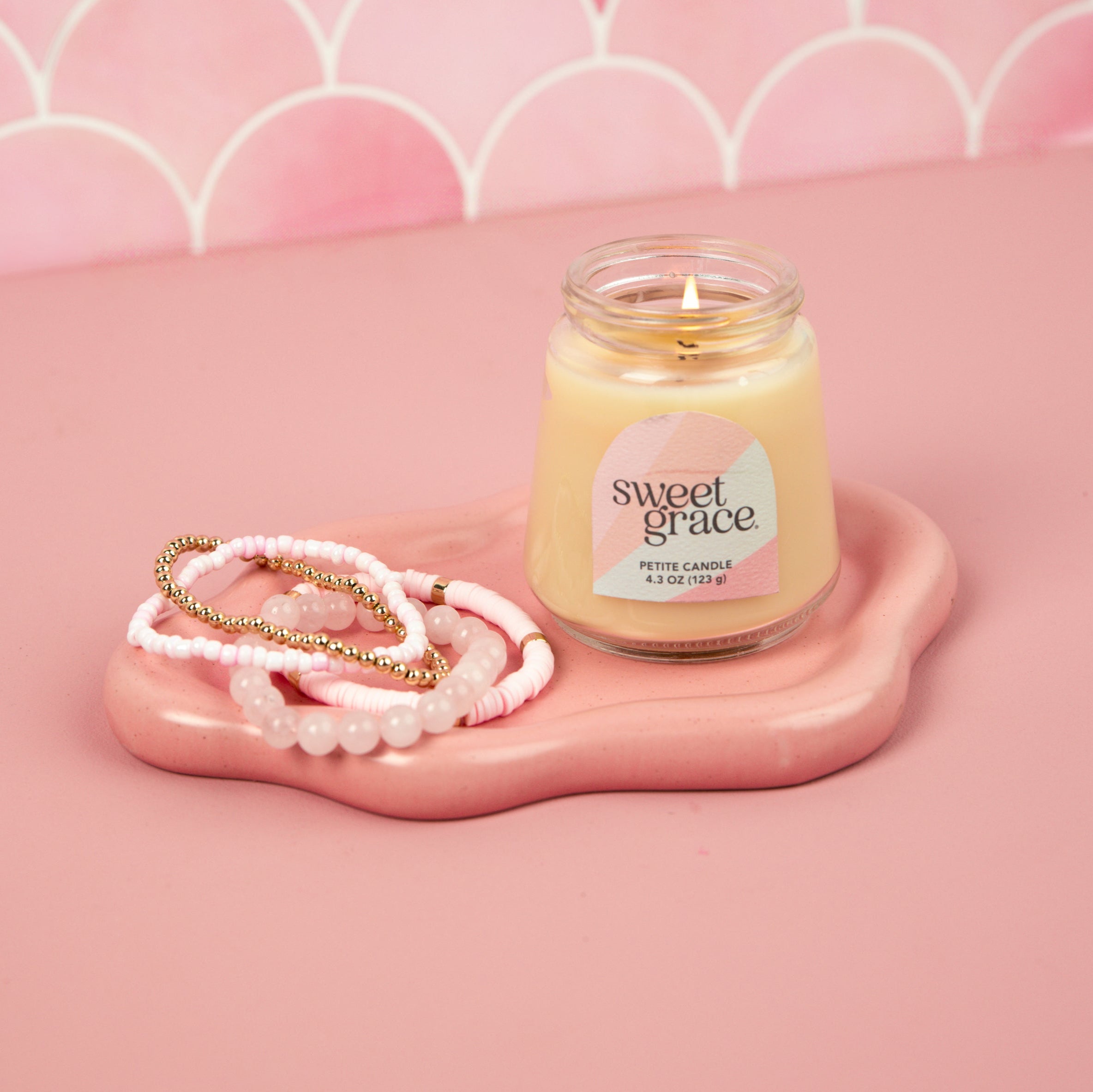 A lit Sweet Grace Collection Candle #078 by Bridgewater Candle in a petite glass jar sits on a pink tray beside three beaded bracelets, set against a pink scalloped background.