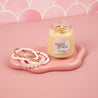 A lit Sweet Grace Collection Candle #078 by Bridgewater Candle in a petite glass jar sits on a pink tray beside three beaded bracelets, set against a pink scalloped background.