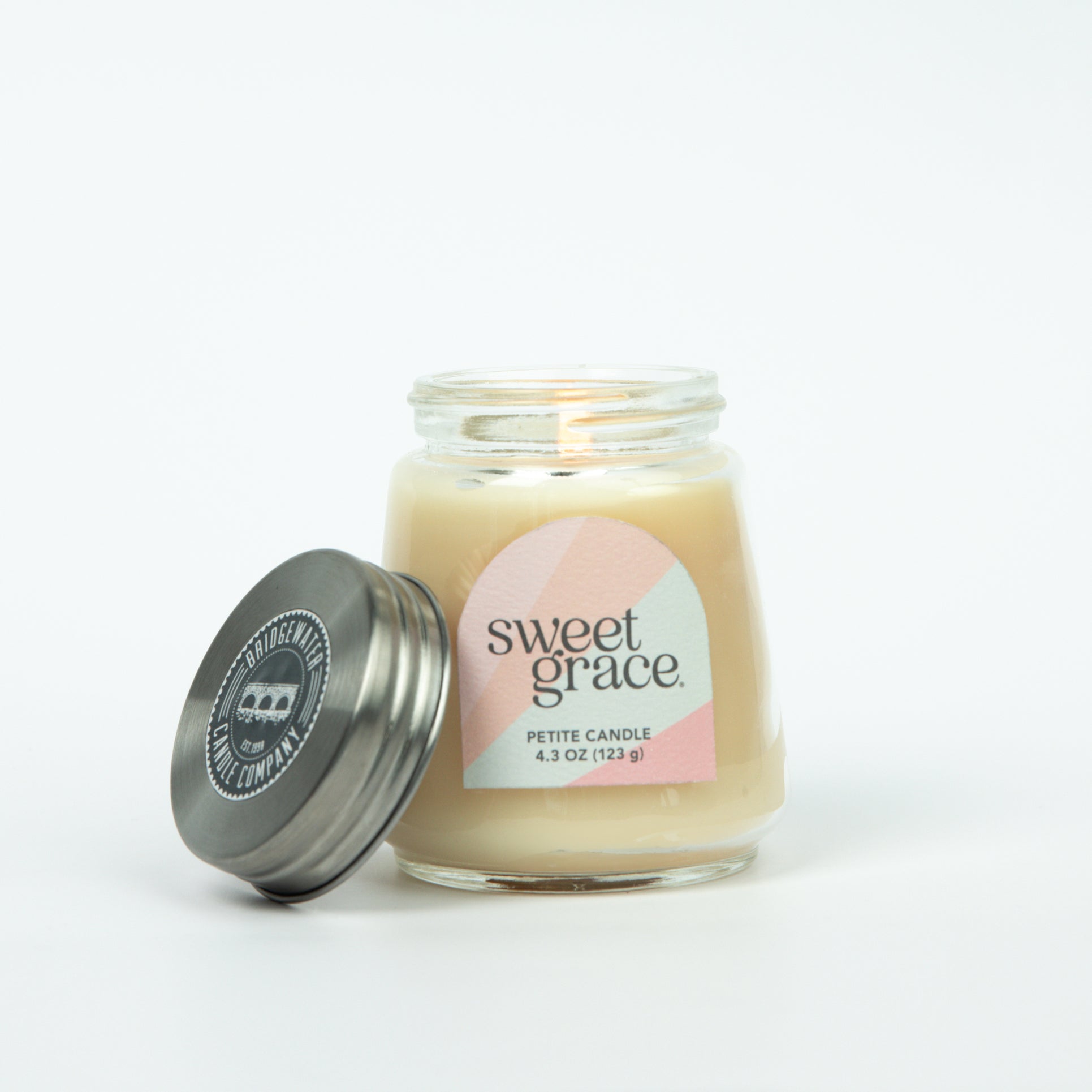 A lit Sweet Grace Collection Candle #078 by Bridgewater Candle in a petite glass jar glows beside its metal lid on a white background. The label shows the scent name and 4.3 oz (123 g) weight of the soy-blend wax.