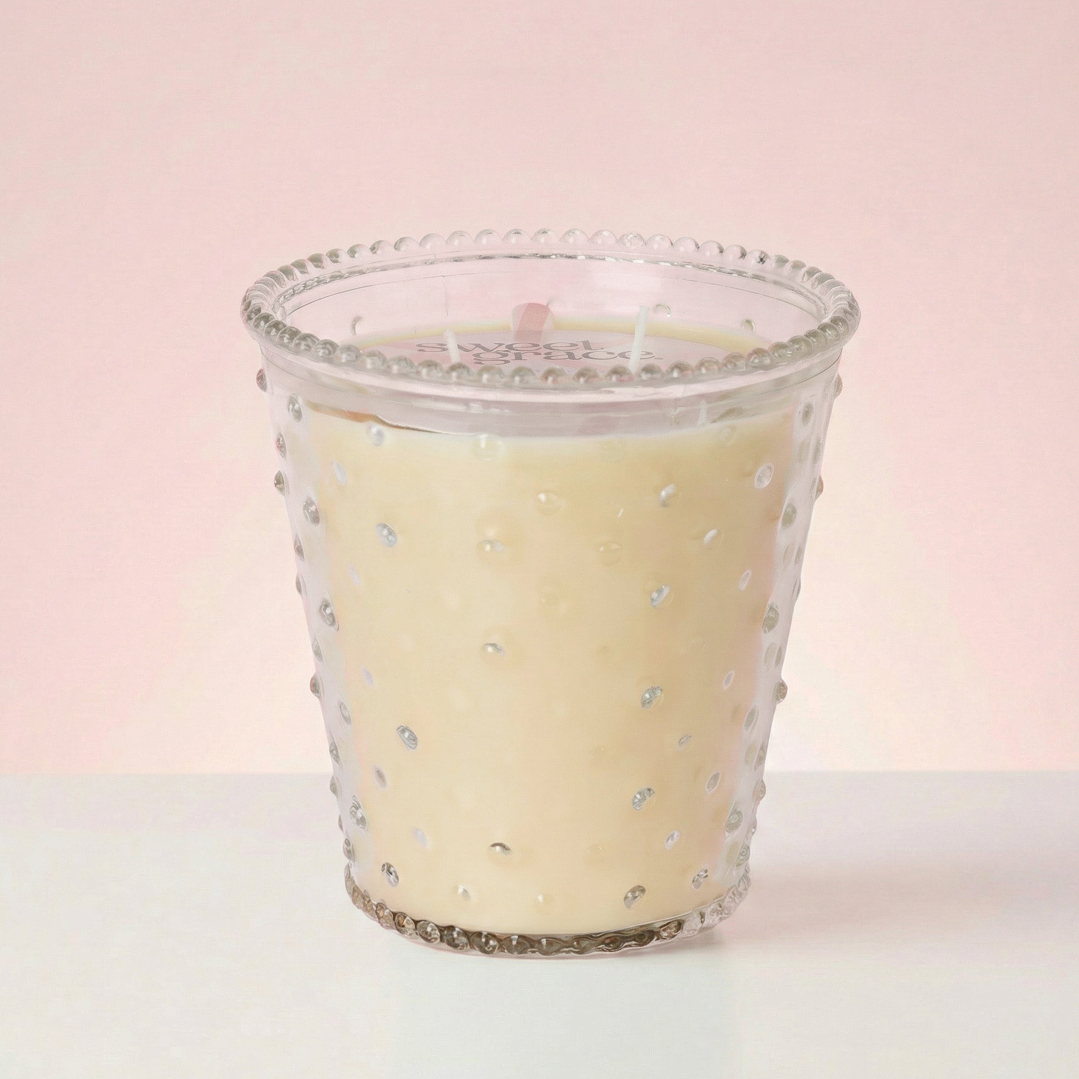 The Bridgewater Candle Sweet Grace Collection Candle #080, featuring a cream hue and three wicks in soy-blend wax, glows in textured hobnail glass atop a white surface with a soft pink background.