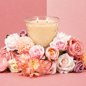 A glowing Sweet Grace Collection Candle #080 by Bridgewater Candle, in a cream decorative glass holder, is surrounded by pink and white flowers against a pink background.