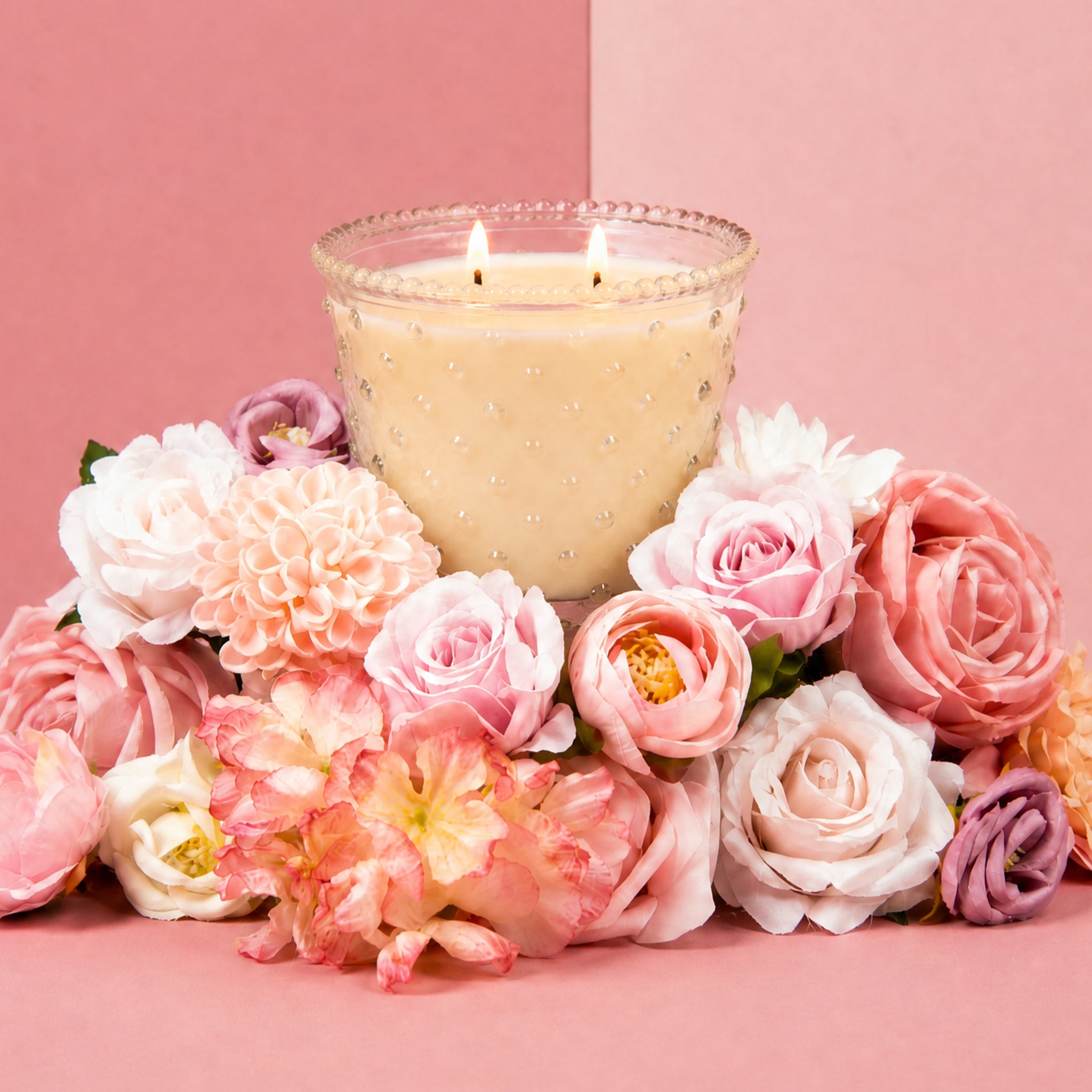 A glowing Sweet Grace Collection Candle #080 by Bridgewater Candle, in a cream decorative glass holder, is surrounded by pink and white flowers against a pink background.