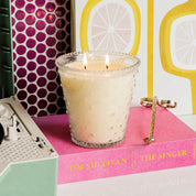 A lit Bridgewater Candle Sweet Grace Collection Candle #080 in a hobnail glass holder sits on the pink book "Jim Sullivan: The Singer," with a gold wick trimmer nearby and colorful, patterned decor in the background.