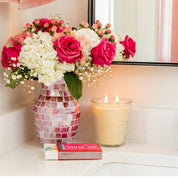 A mosaic vase with pink and white flowers sits on a bathroom counter next to a lit Bridgewater Candle Sweet Grace Collection Candle #080 and a colorful STRIKING box, with their reflection visible in the mirror.