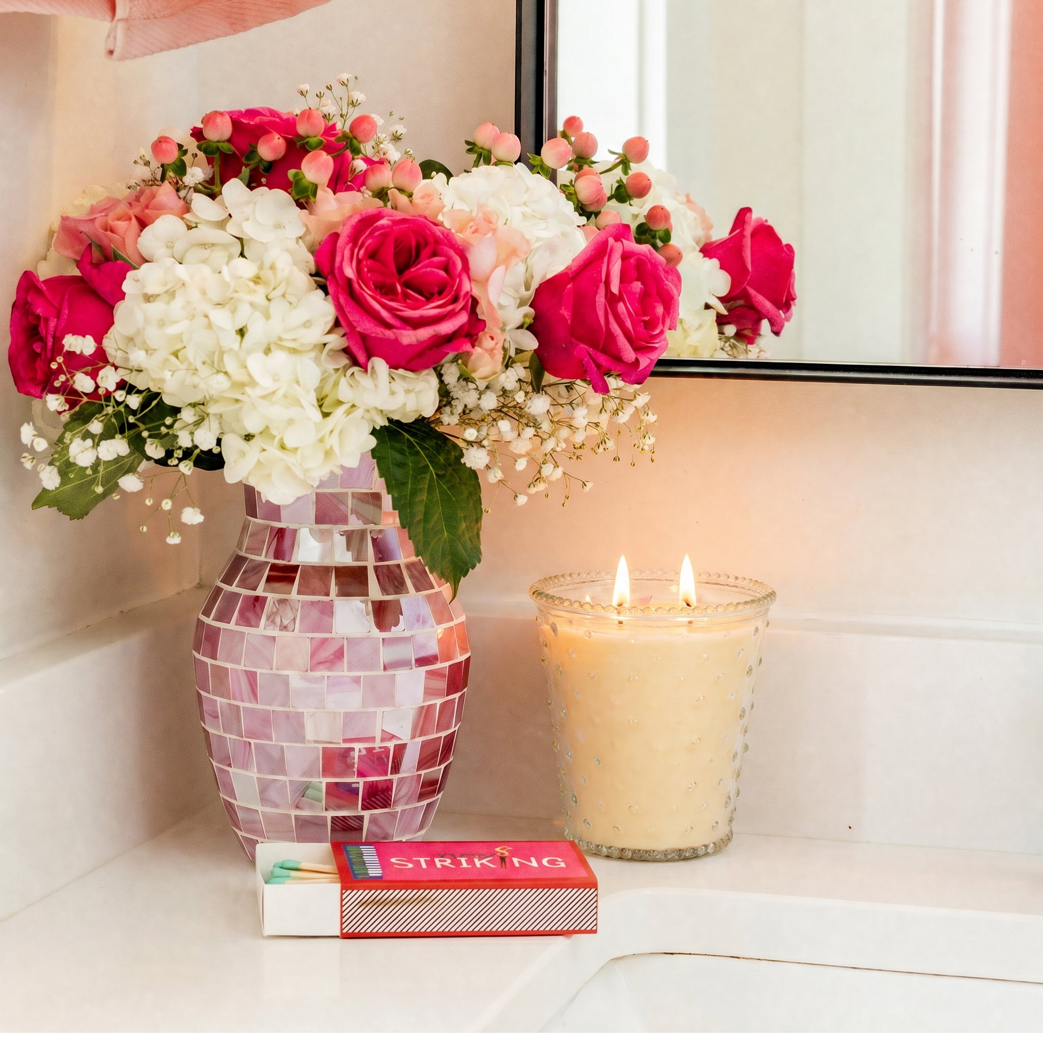 A mosaic vase with pink and white flowers sits on a bathroom counter next to a lit Bridgewater Candle Sweet Grace Collection Candle #080 and a colorful STRIKING box, with their reflection visible in the mirror.