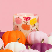 The Bridgewater Candle Sweet Grace Pumpkin Decal Candle, a soy candle in a glass holder adorned with vibrant pumpkin illustrations, is surrounded by small artificial pumpkins in orange, pink, purple, and white and set against a pink background.