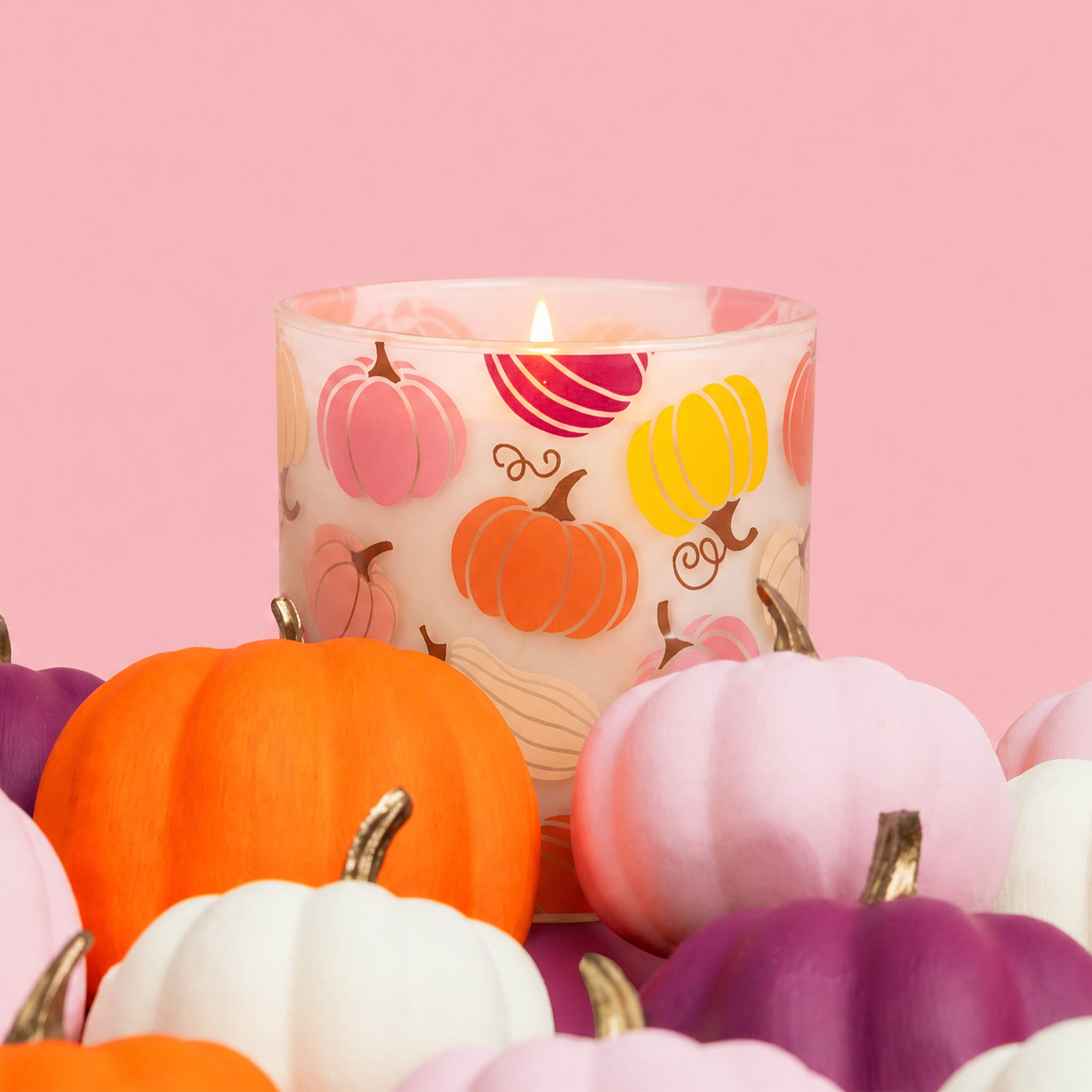 The Bridgewater Candle Sweet Grace Pumpkin Decal Candle, a soy candle in a glass holder adorned with vibrant pumpkin illustrations, is surrounded by small artificial pumpkins in orange, pink, purple, and white and set against a pink background.