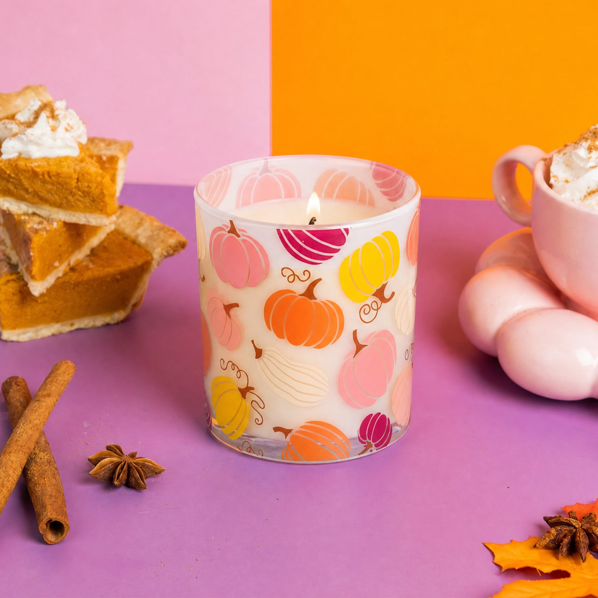 The Bridgewater Candle Sweet Grace Pumpkin Decal Candle, featuring a glass holder adorned with colorful pumpkin designs, rests on a purple surface among cinnamon sticks, star anise, slices of pumpkin pie, and a pink cup, set against an orange and pink background.