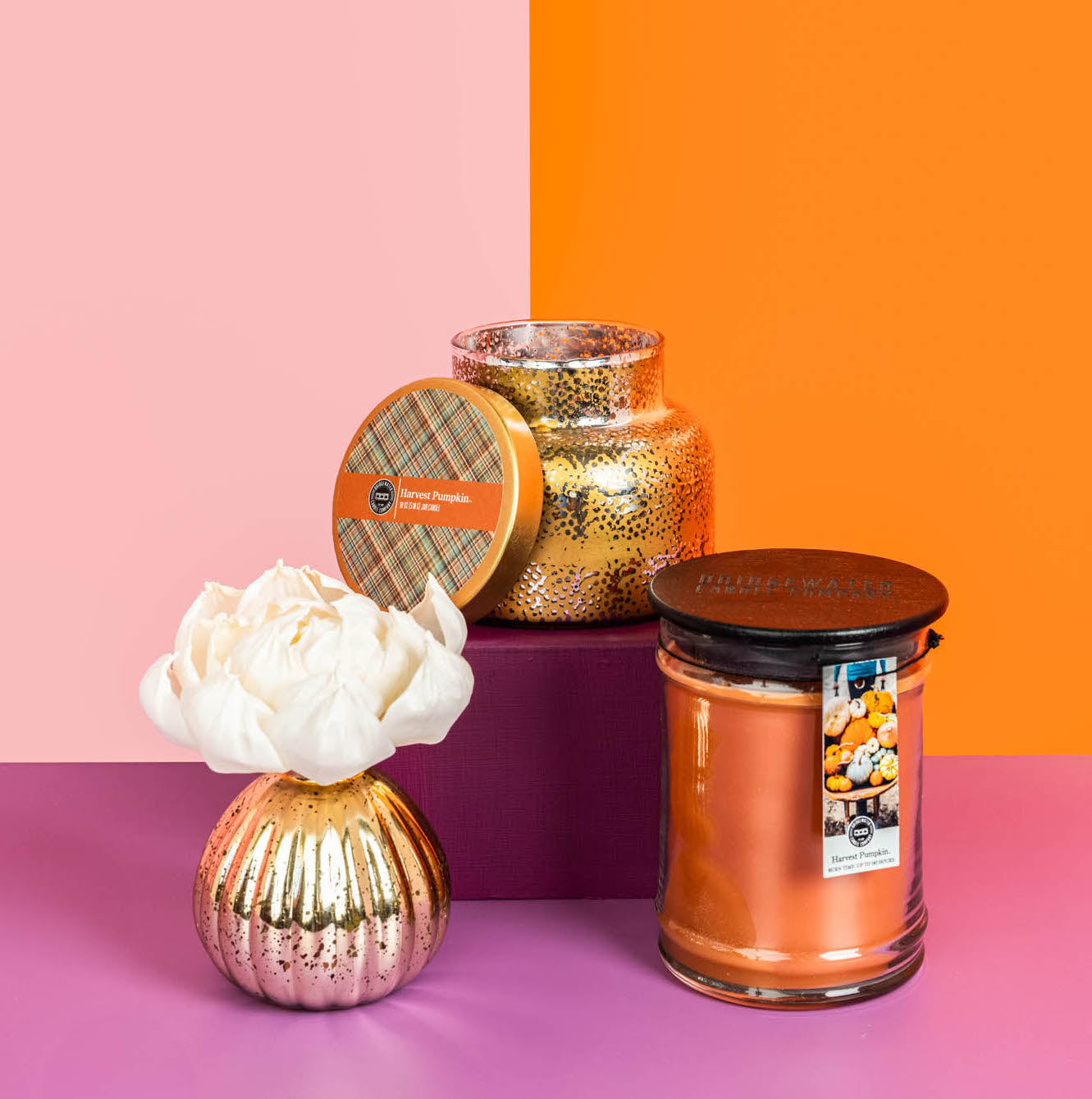 Three decorative candles sit on a purple box: a Bridgewater Candle Company Harvest Pumpkin 18oz Large Jar Candle, a gold candle with metallic lid, and a gold vase holding a white flower. The background is split orange and pink.
