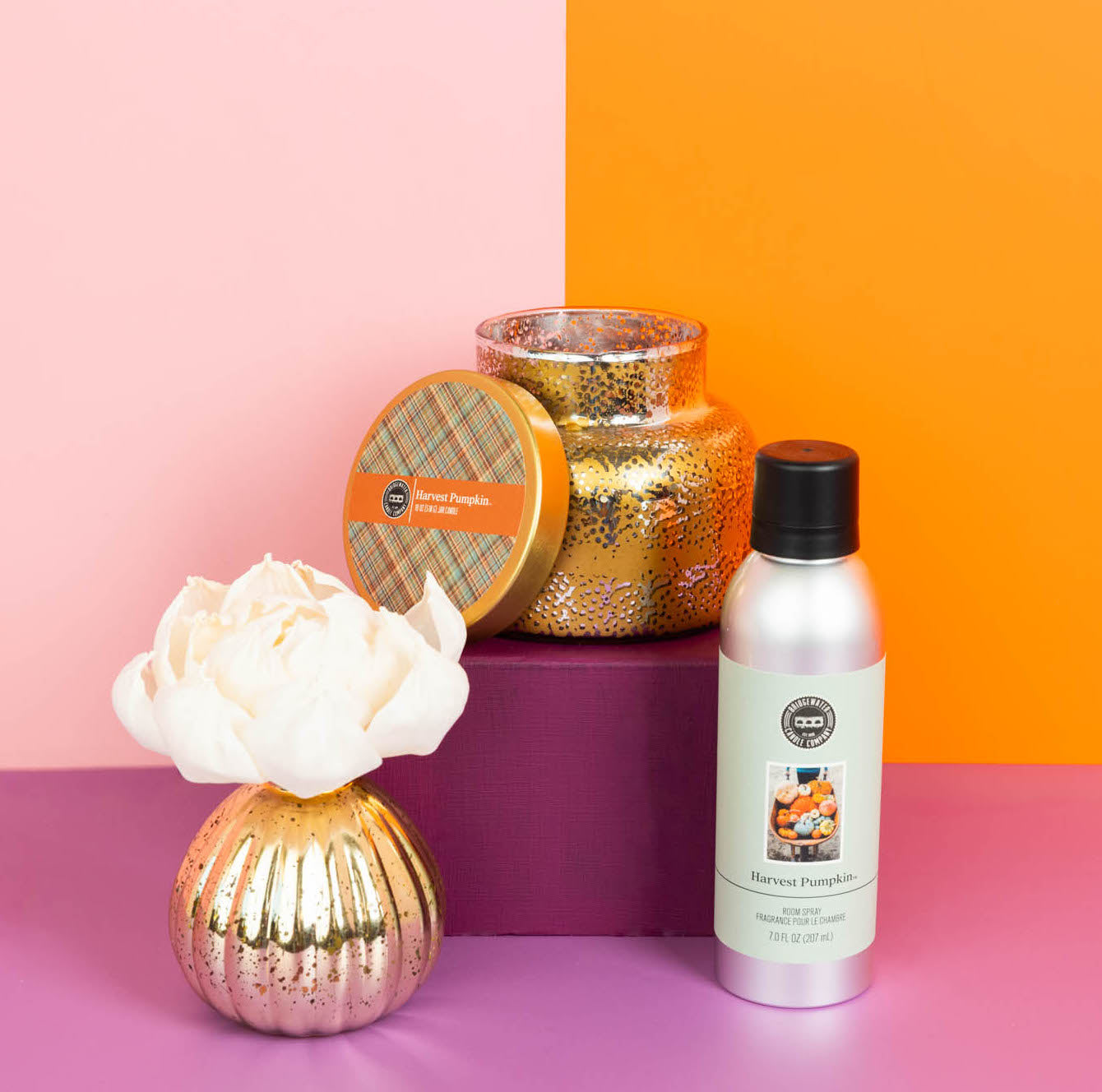 Three harvest pumpkin-themed products—a scented candle with lid, a silver fragrance spray, and the Bridgewater Candle Company Harvest Pumpkin Mercury Flower Diffuser—are arranged on a purple block against a pink and orange background. Enjoy the inviting aroma of pumpkin spice fragrance as you bring seasonal charm to any space.